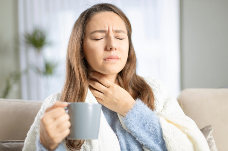 A woman holds her sore throat while holding a cup of green tea in the other hand