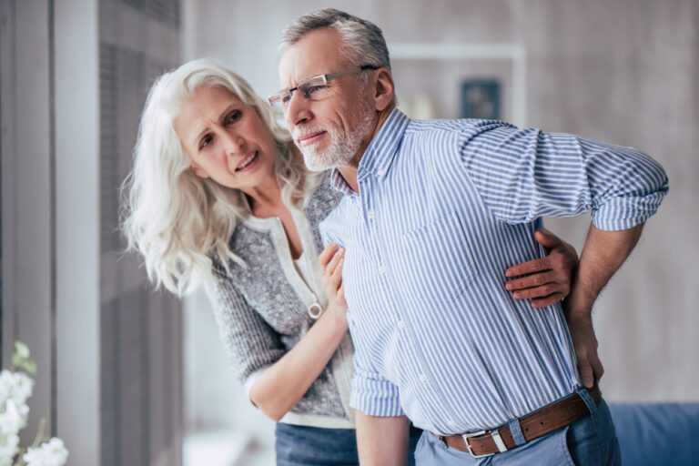 A man in his 60s holds his lower back in pain while being supported by a woman of a similar age.