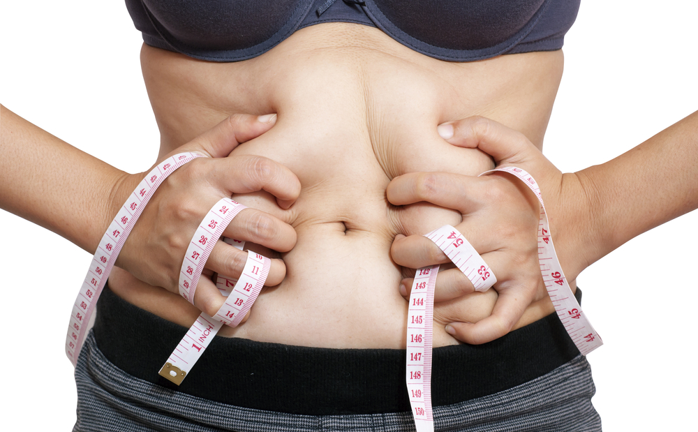 A woman tugs at her stomach while holding a tape measure, frustrated with her weight loss plateau.