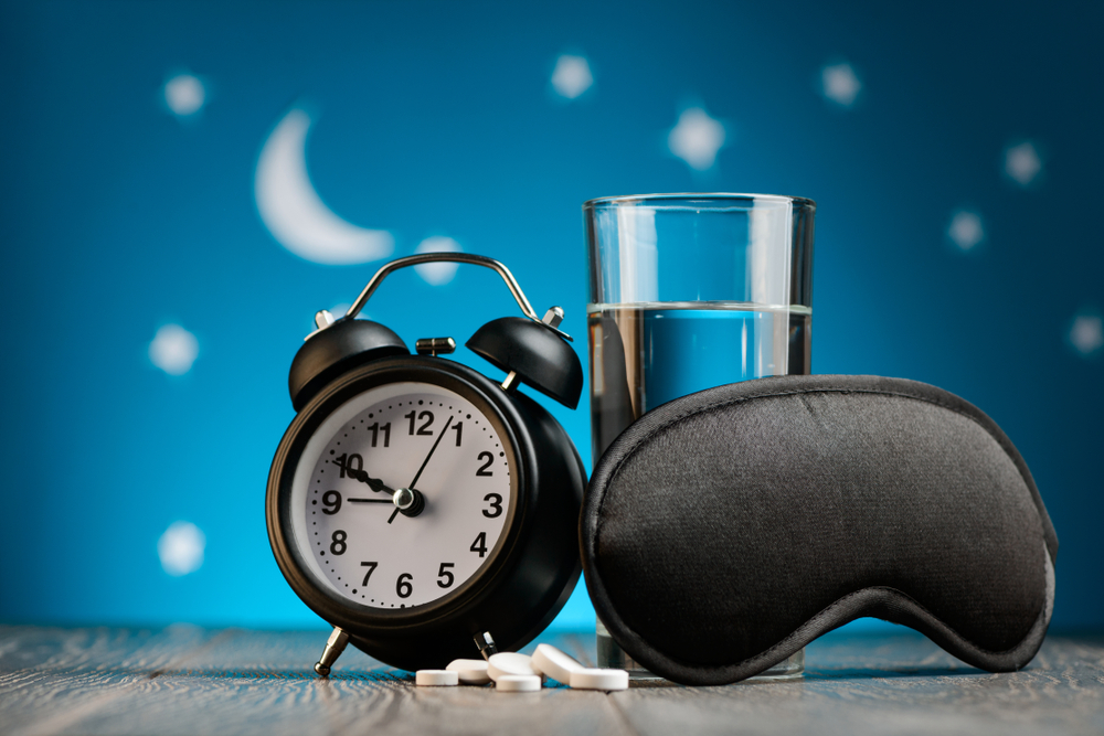 A photo of a sleeping mask, alarm clock, a glass of water and melatonin tablets against a backdrop of stars.