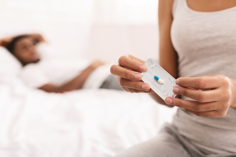 A woman sits on the side of her bed with her partner in the background, she is holding the morning after pill in her hand.