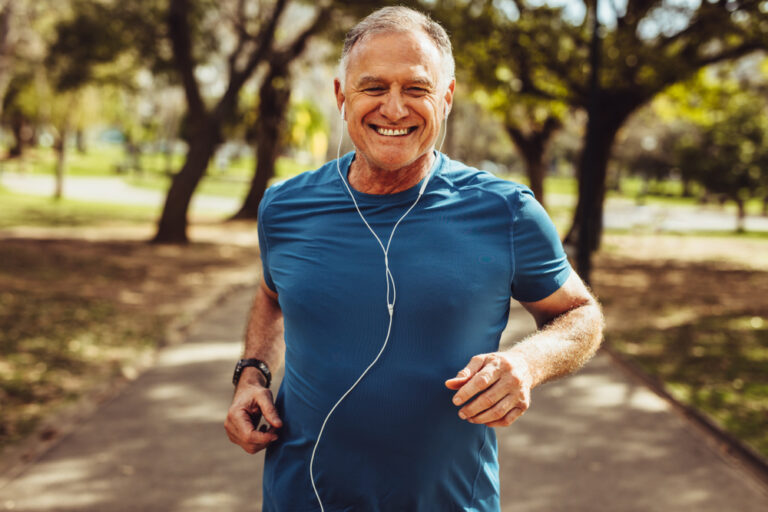 A man in his 60s is running in the park while listening to music. He has a big grin because he's been successful in losing weight.