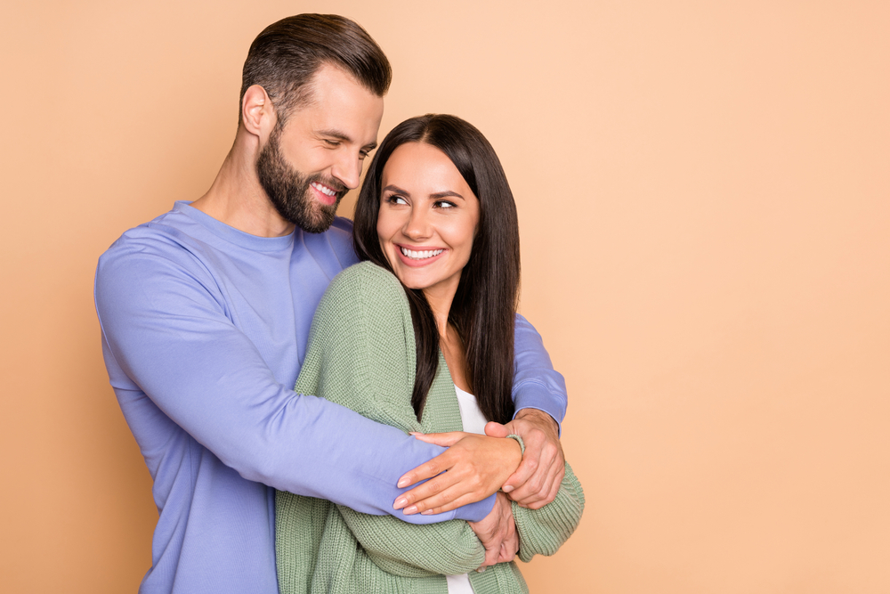 A young couple stand in front of an orange background, hugging, with great hair.