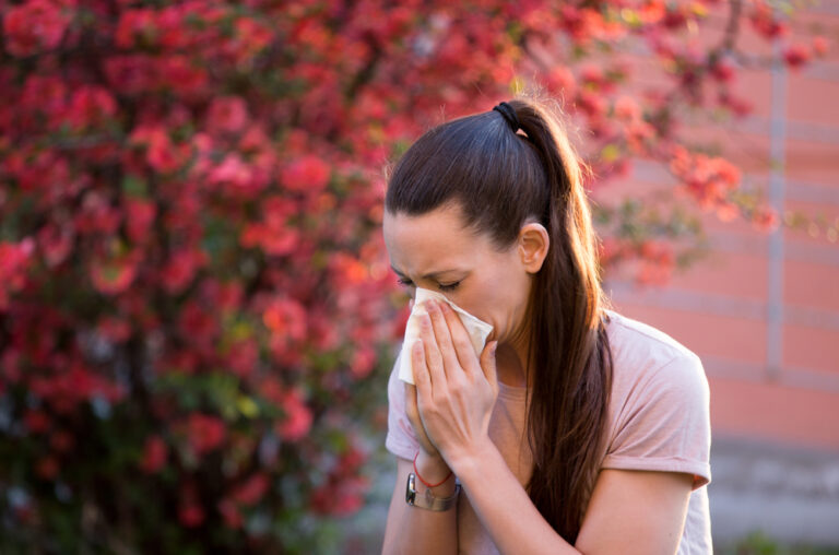 A woman with hay fever sneezes into a tissue while stood in front of a tree with pink blossom