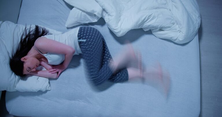 A topdown photo of a woman laying on her bed with restless legs syndrome, her legs are blurred to show the constant movement as she tries to sleep.