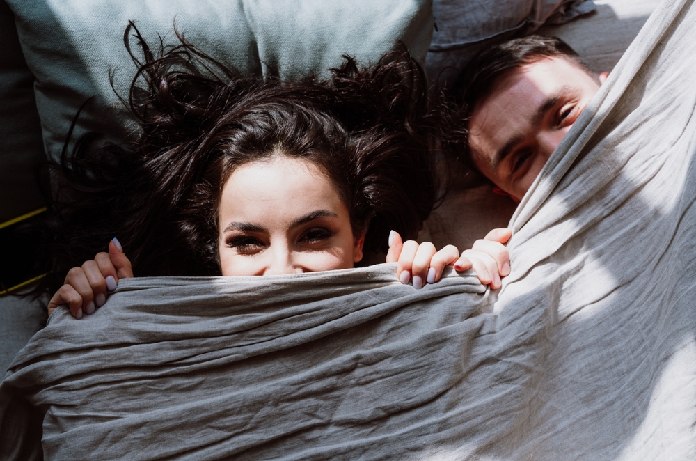 A young couple happily peek out from under their bed sheets after using sildenafil to combat his erectile dysfunction.