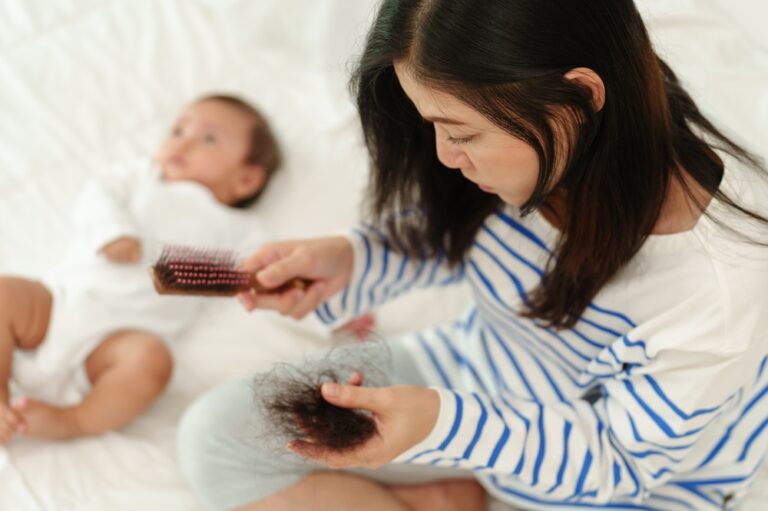 A young mother has a ball of hair in her hand after brushing her hair, with her baby on the bed in the background.