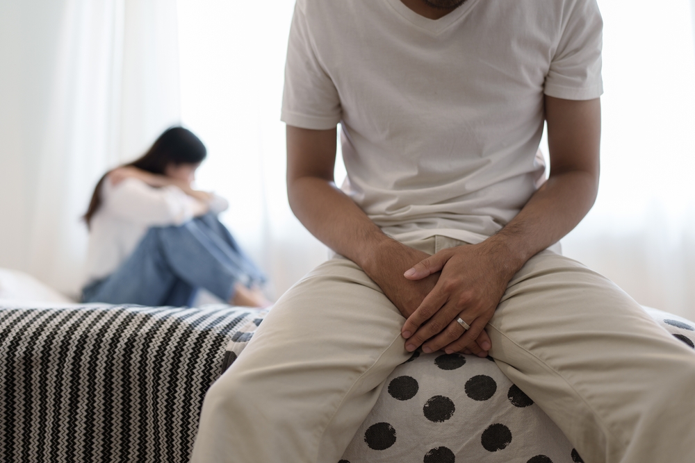 A man sits on the edge of the bed with his hands on the outside of his jeans in his genital area due to erectile dysfunction, while his girlfriend sits in the background looking upset with the situation.