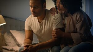 A woman comforts her boyfriend at the edge of the bed, after dealing with erectile dysfunction.