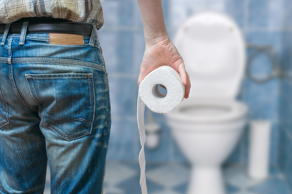 A man stands in front of the toilet holding a roll of toilet paper, anticipating diarrhoea