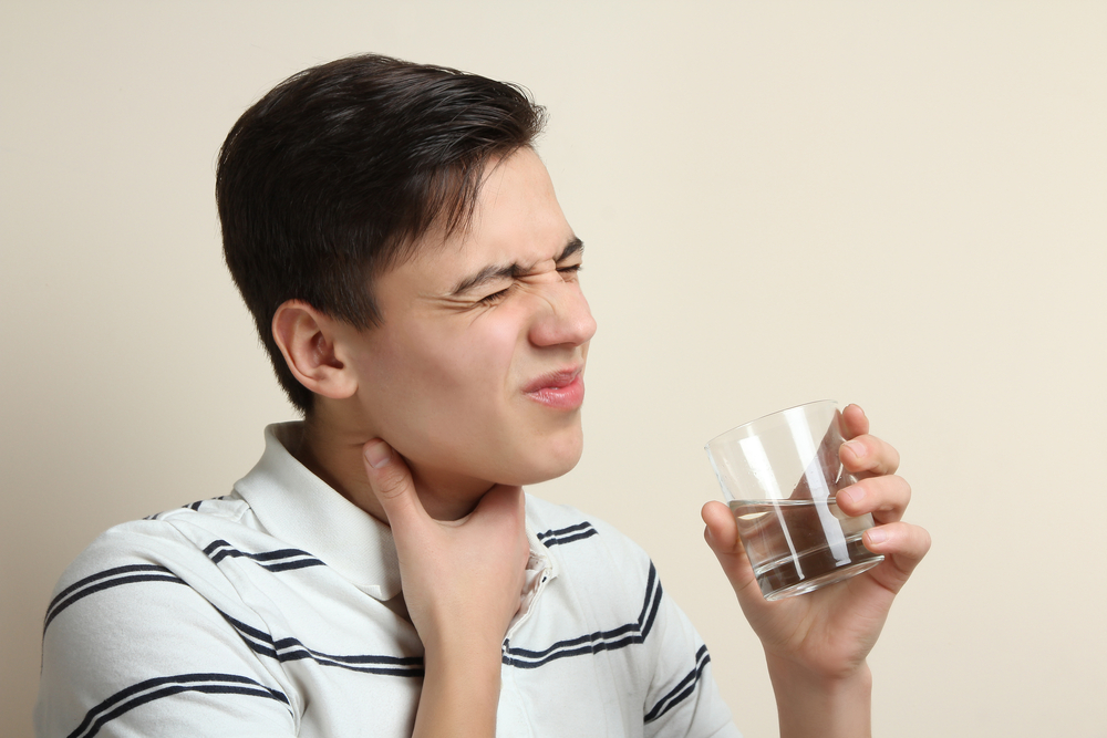 A young man is holding his throat in pain after a cough, he is holding a glass of water.