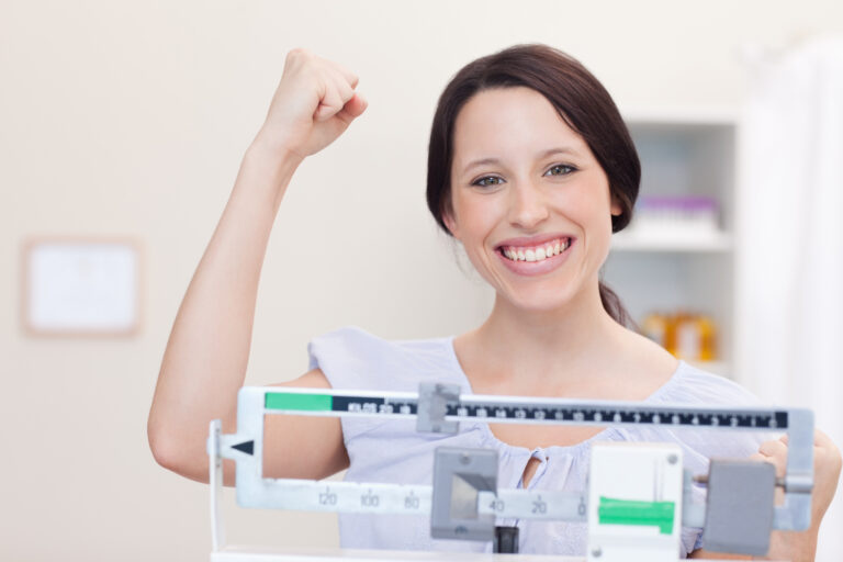 A woman pumps her fist in excitement while stood on a scale, she's happy with the progress she's made in her weight loss journey.